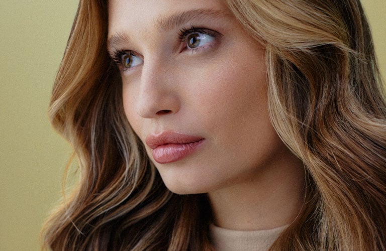 Foto de una mujer con cabello lacio, largo y rubio, peinado hacia un lado. El fondo es amarillo claro, y ella lleva una blusa negra de un solo hombro.